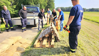 Schulungsteilnehmer bauen eine Deicherhöhung mit Europaletten. Im Hintergrund steht ein Pritschenwagen, am Rand der Deichkrone liegt eine Europalette auf dem Boden, zwei weitere stehen aneinandergelehnt davor und darüber. Seminarteilnehmer und Ausbilder tragen weitere Europaletten herbei. Das Wetter ist sonnig. 