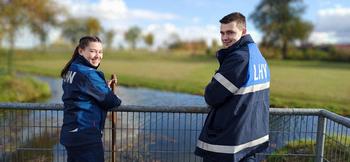 Zwei Jugendliche mit blauen LHW-Jacken stehen an einem Geländer vor einem Gewässer und lächeln in die Kamera. 