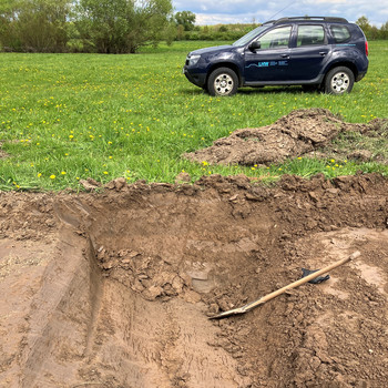 Deichbau Booser Bogen: Bevor die eigentlichen Bauarbeiten beginnen konnten, haben die Archäologen das Baufeld untersucht. Auf dem Bild ist im Hintergrund ein Fahrzeug des LHW auf einer begrünten Fläche zu sehen. Im Vordergrund ist eine Grube aus der Erde ausgehoben worden. Darin liegt eine Schaufel.